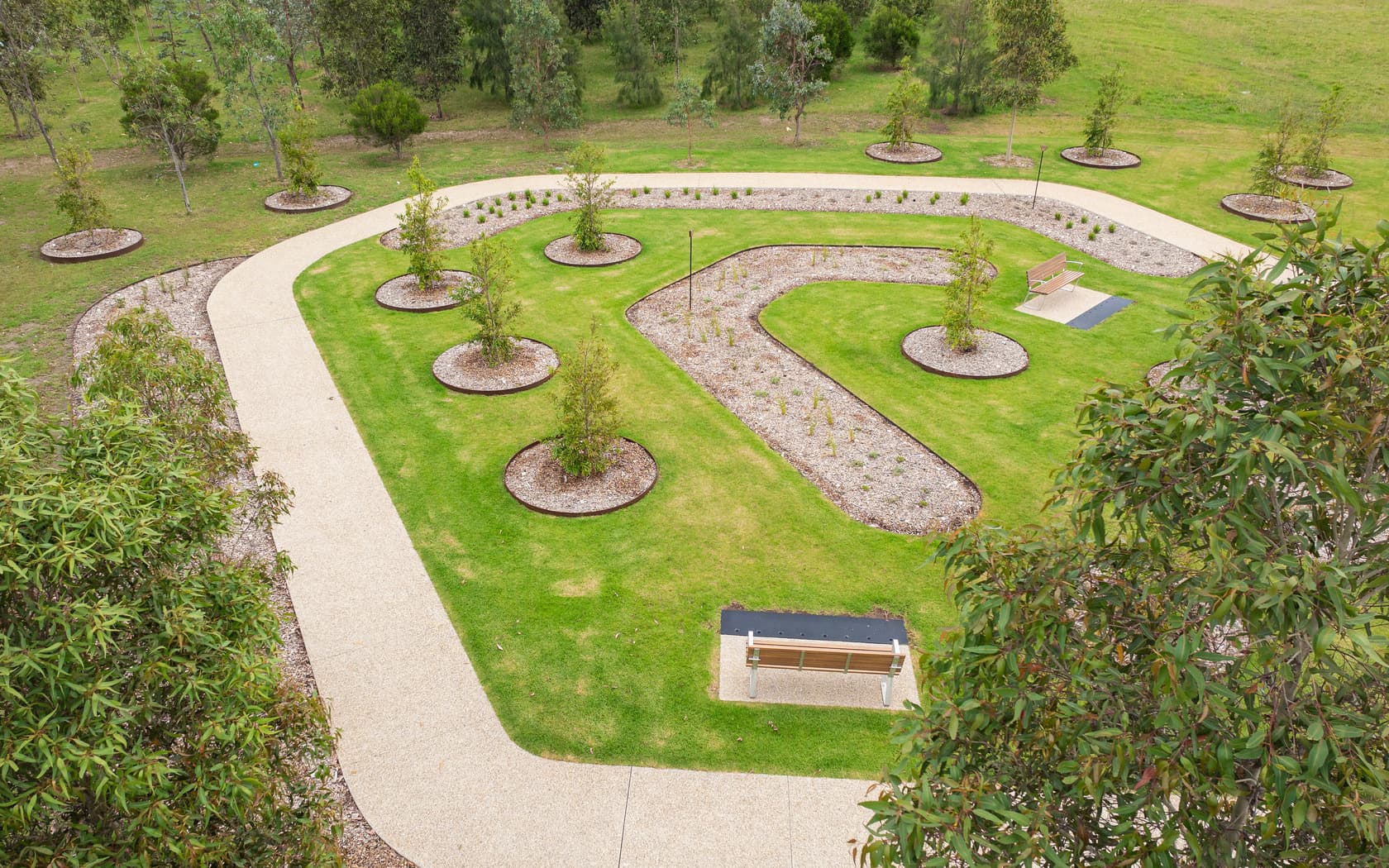 Aerial view of a landscaped park featuring curved pathways, circular flower beds, small trees, and a bench on a grassy lawn.