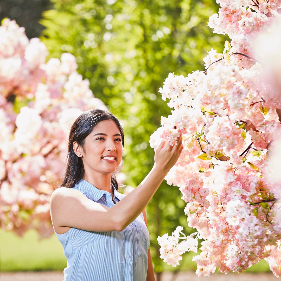Woman in a sleeveless blouse gently touches pink cherry blossoms in a sunlit garden.