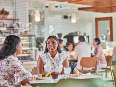 A mother and daughter sit at Cafe Vita et flores in Bunurong Memorial park. They have sandwiches and wraps on the plates in front of them. The daughter is smiling and the mother has the back of her head to camera.