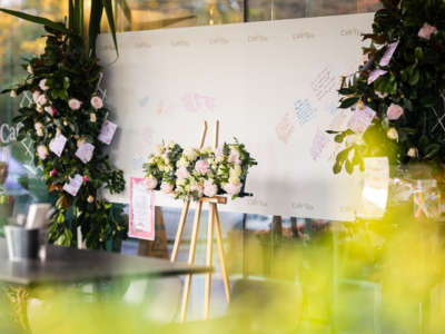 A large, cream-coloured backdrop features messages scrawled on them in different coloured ink. Pink and white floral arrangements on the left and right frame the backdrop.