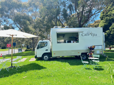 The Cafe Vita et flores food truck is parked on a green lawn, beneath trees on a sunny day. An umbrella is set up to the left-hand side, and some chairs are set up in front of the truck on the right.