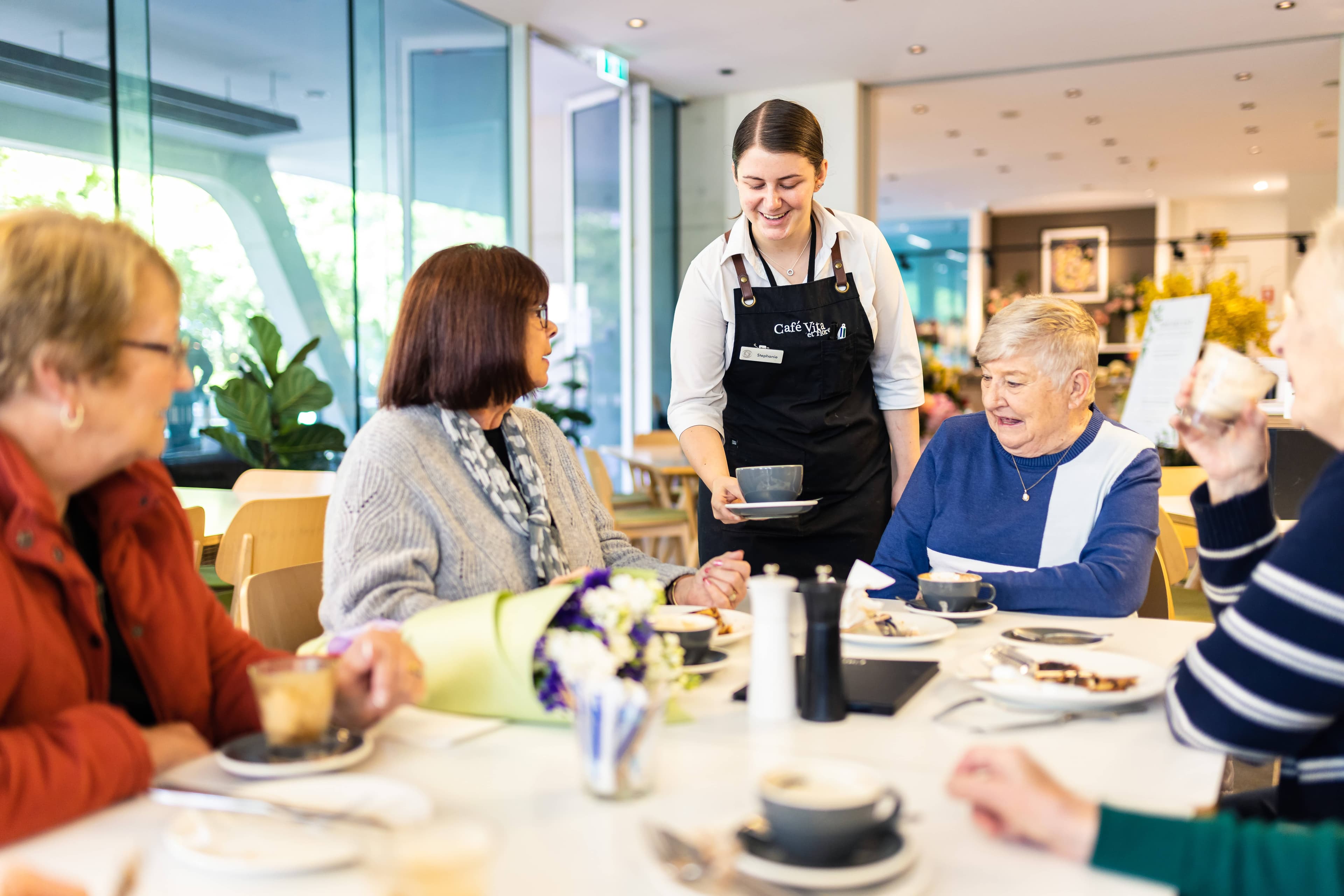 Friendly café staff serving freshly brewed coffee to smiling customers in a cozy, welcoming café setting
