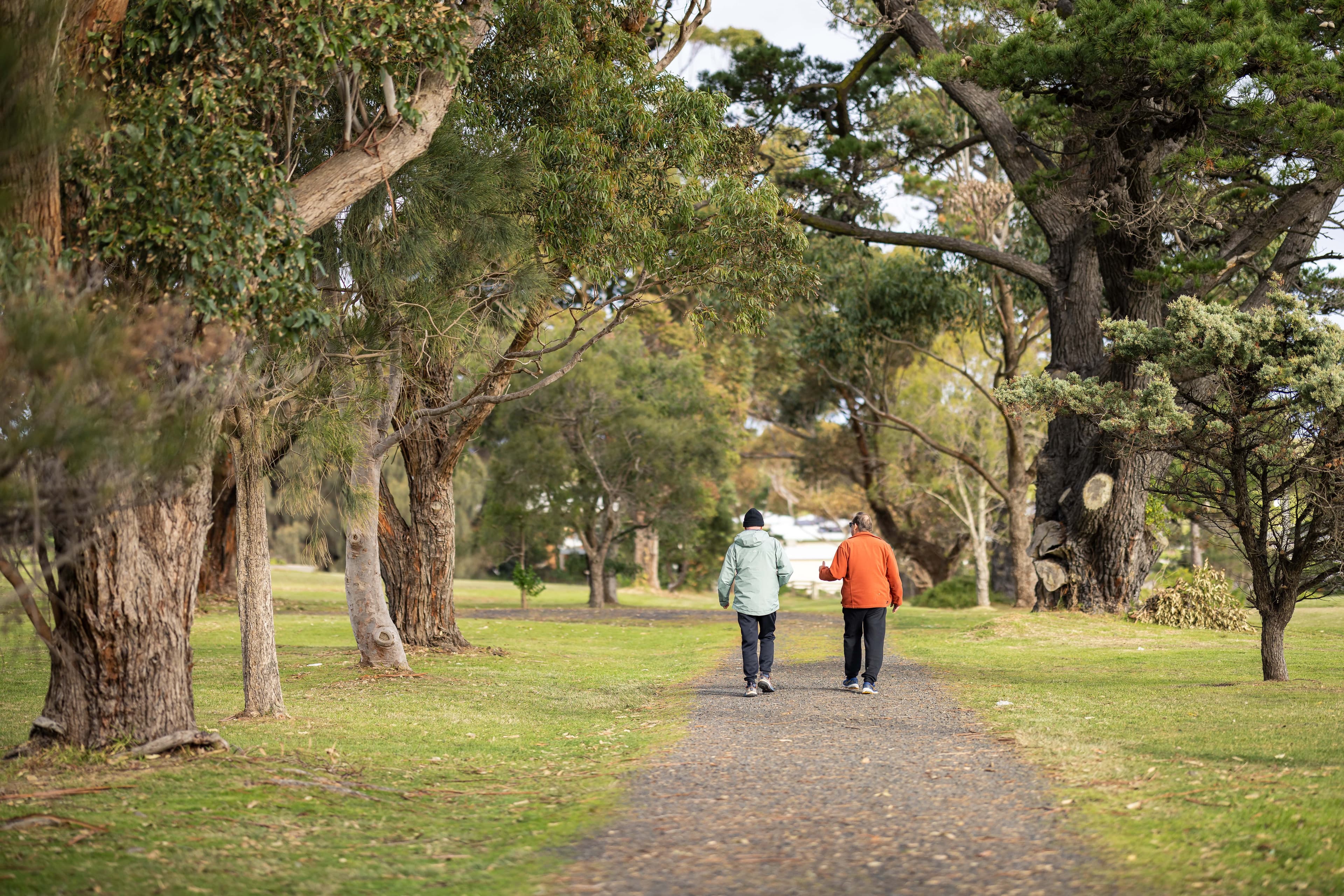 A couple walking away into the distance along a gravel path, surrounded by lush green trees in a peaceful, natural setting.